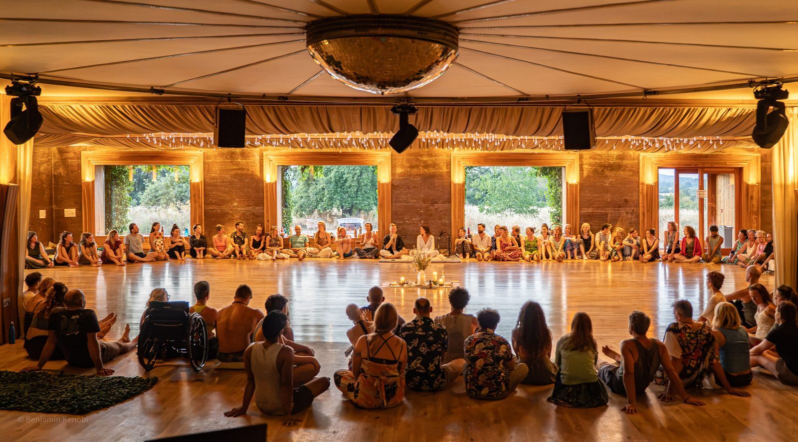 A large circle of people, including a wheelchair user, sits together on the Gillyflower floor around a candlelit centrepiece.