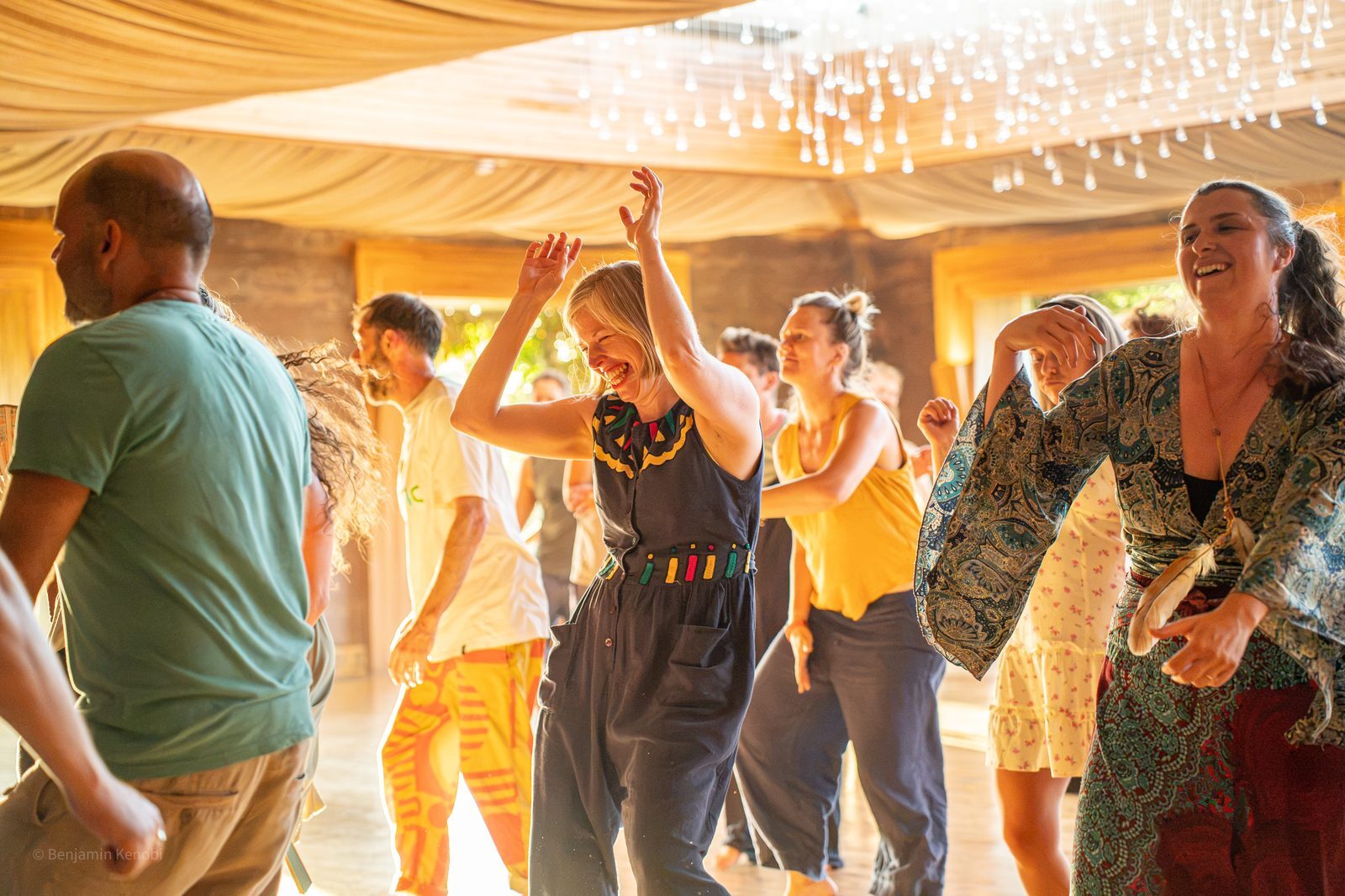 A woman laughs with her arms raised as people dance freely around her in warm evening light.