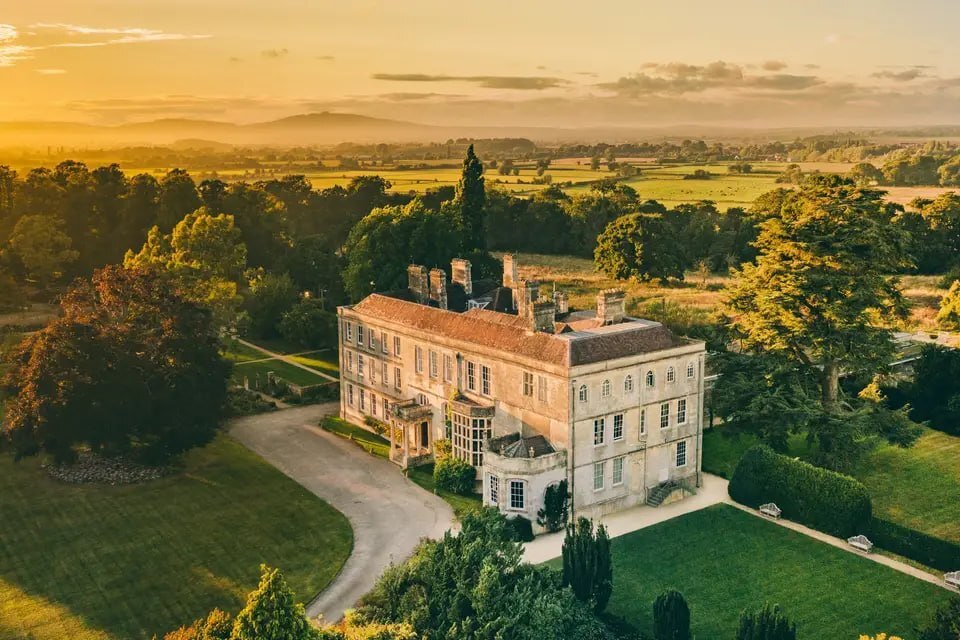 Aerial view of Elmore Court's stone manor at sunset, surrounded by ancient trees and golden fields.